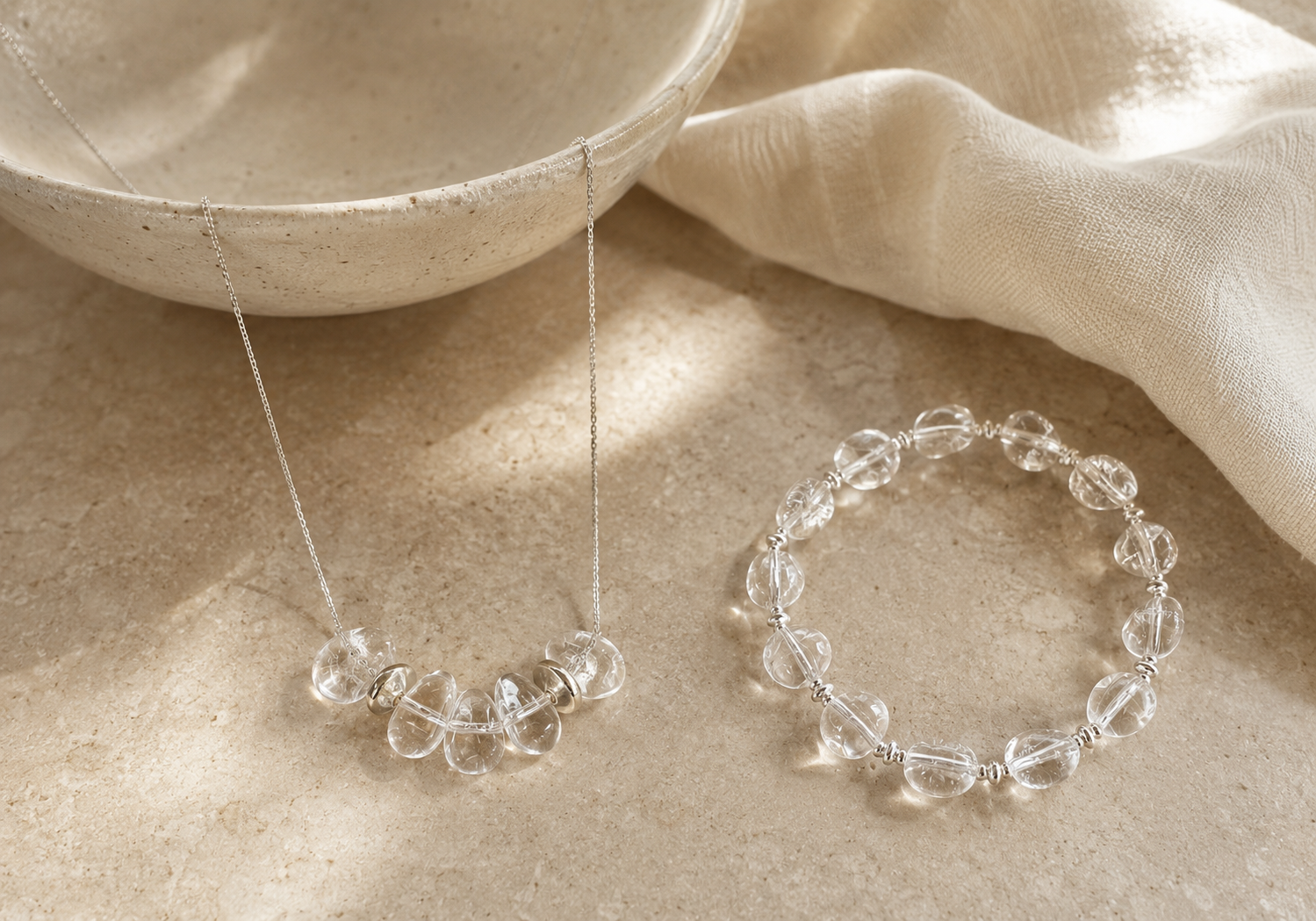 Clear quartz necklace and bracelet styled with a ceramic bowl, soft ivory fabric, and warm stone textures.