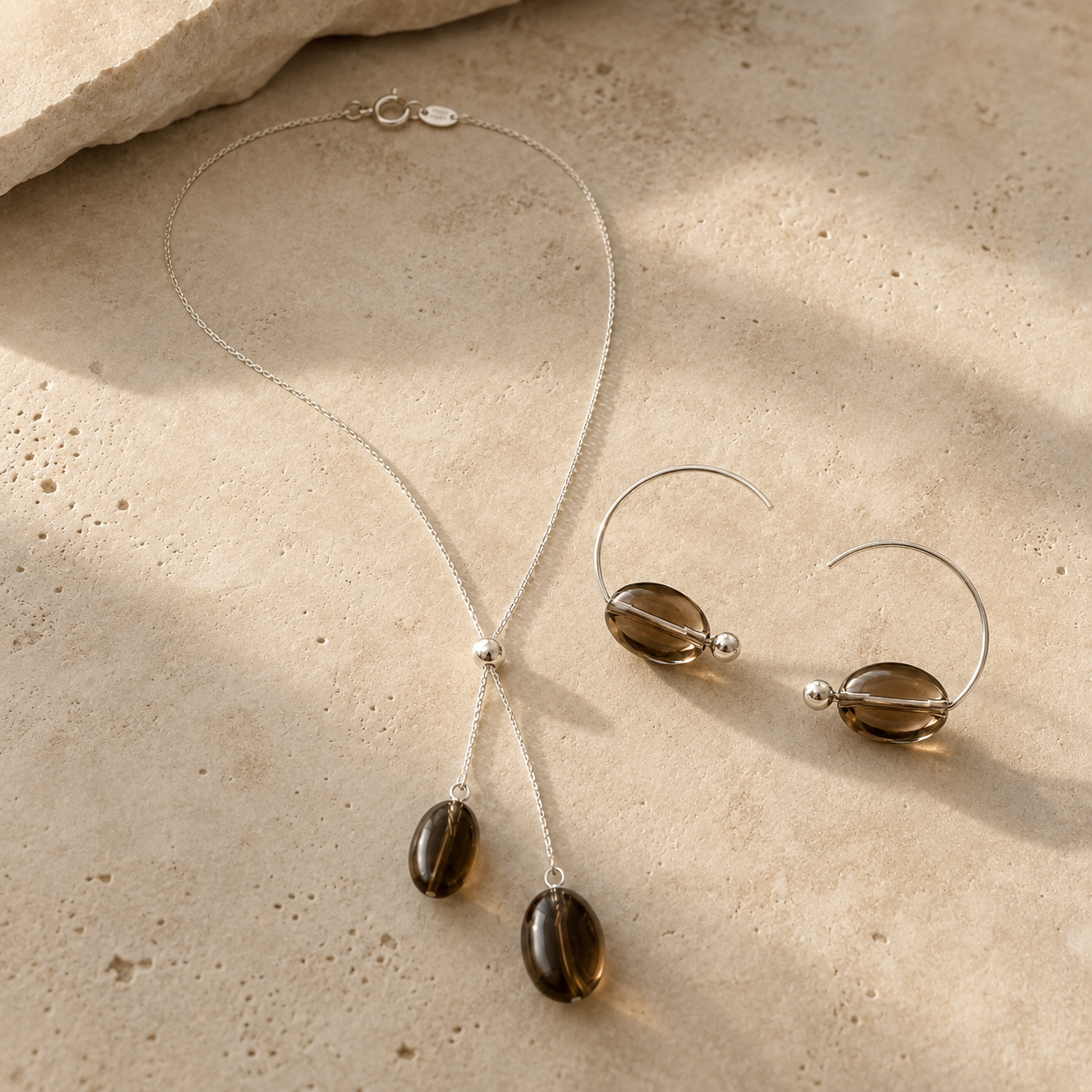 Smoky quartz necklace and earrings arranged on warm natural stone for the LUNARAE Grounding collection.