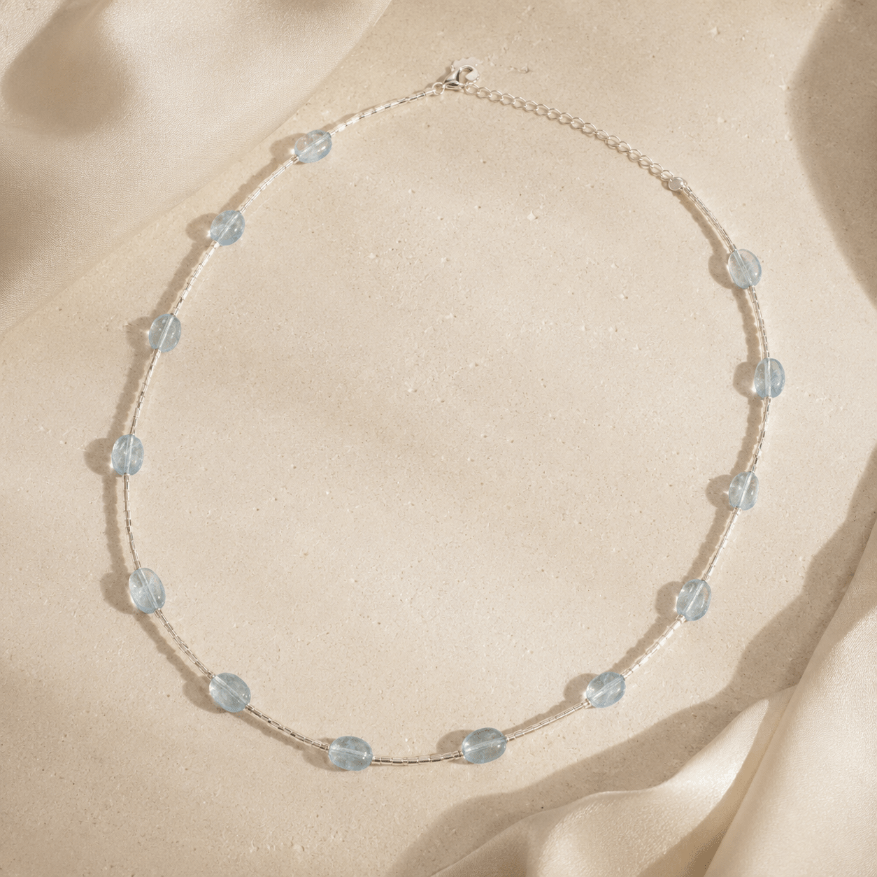 Aquamarine Tube Necklace styled on a warm stone surface with softly draped ivory satin and natural sunlight