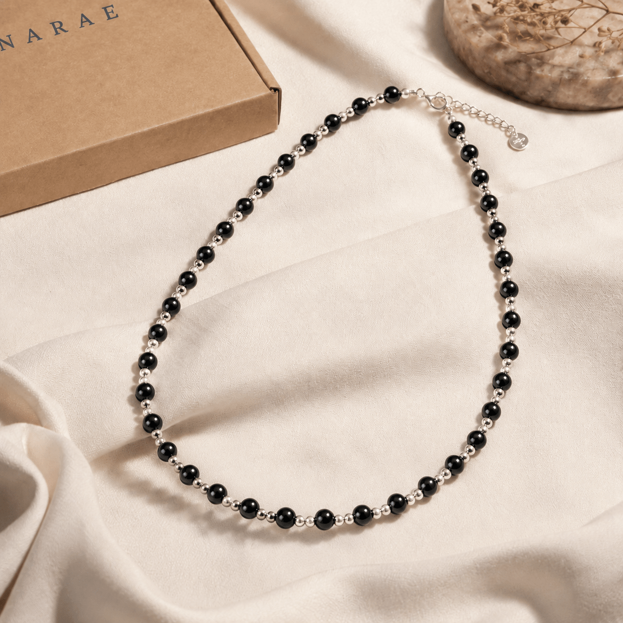 Black Onyx Bead Necklace styled on softly draped ivory fabric beside LUNARAE packaging and a neutral stone accent