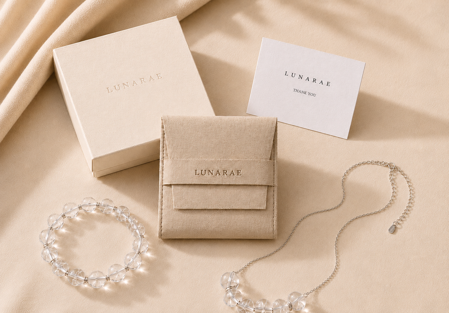 LUNARAE clear quartz bracelet and necklace displayed with a soft jewelry pouch, gift box, and thank you card on a warm beige background.