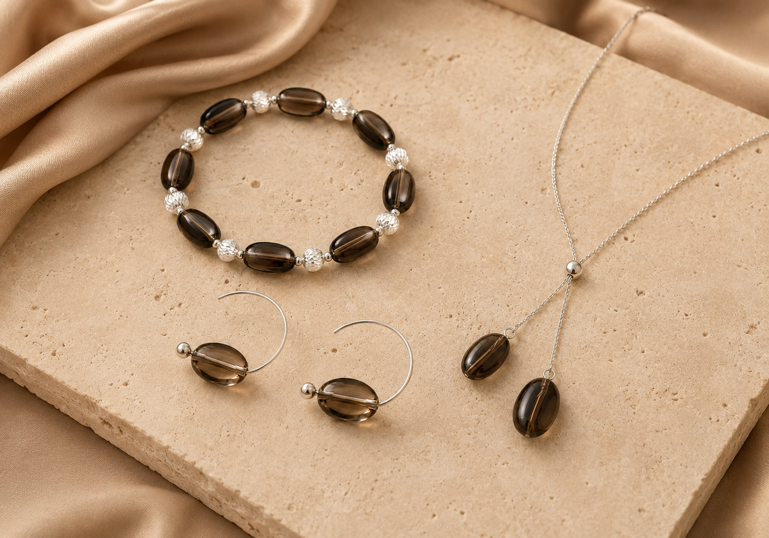Smoky quartz bracelet, Y necklace, and hoop earrings from the LUNARAE Grounding Collection styled on warm beige stone and satin.
