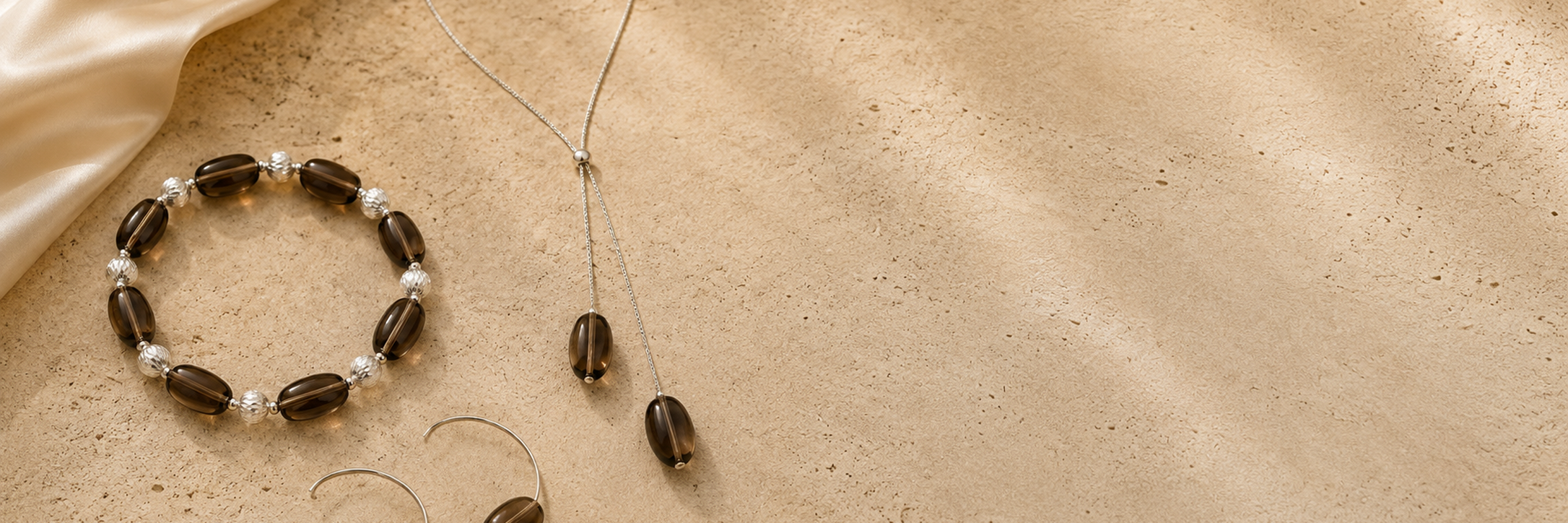 Smoky quartz Y necklace, stretch bracelet, and open hoop earrings styled on warm travertine with soft ivory satin.