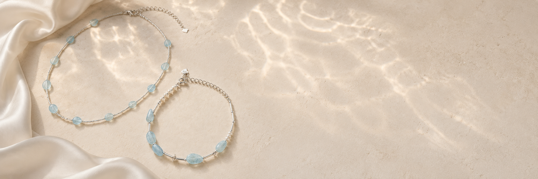 Aquamarine tube necklace and aquamarine pearl bracelet styled on warm stone with soft ivory satin and water reflections.